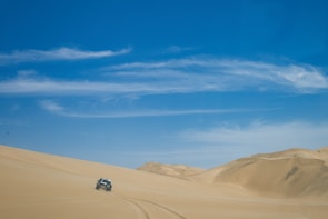 Expansive golden sand dunes of Al Ula desert under a clear blue sky, with a luxury SUV driving along a winding road.
