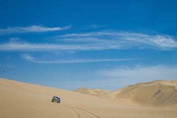 A vast, sandy desert landscape under a clear, blue sky. A four-wheel-drive vehicle is seen navigating the dunes, leaving tracks in the sand behind it. The dunes are gently curving and stretch towards the horizon.