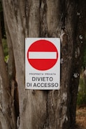A weathered tree trunk serves as the background for a metal sign displaying a red circle with a white horizontal bar inside it. Beneath the circle, the text reads 'PROPRIETA' PRIVATA DIVIETO DI ACCESSO' indicating private property and no entry.