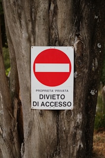 A weathered tree trunk serves as the background for a metal sign displaying a red circle with a white horizontal bar inside it. Beneath the circle, the text reads 'PROPRIETA' PRIVATA DIVIETO DI ACCESSO' indicating private property and no entry.