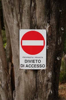 A weathered tree trunk serves as the background for a metal sign displaying a red circle with a white horizontal bar inside it. Beneath the circle, the text reads 'PROPRIETA' PRIVATA DIVIETO DI ACCESSO' indicating private property and no entry.