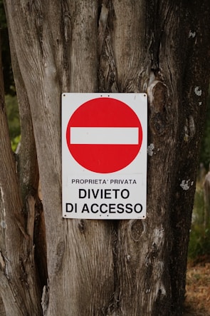 A weathered tree trunk serves as the background for a metal sign displaying a red circle with a white horizontal bar inside it. Beneath the circle, the text reads 'PROPRIETA' PRIVATA DIVIETO DI ACCESSO' indicating private property and no entry.