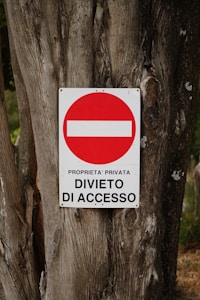 A weathered tree trunk serves as the background for a metal sign displaying a red circle with a white horizontal bar inside it. Beneath the circle, the text reads 'PROPRIETA' PRIVATA DIVIETO DI ACCESSO' indicating private property and no entry.