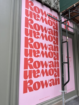 A large pink poster with the word 'Rowan' printed repeatedly in bold red letters. The poster is affixed to an outdoor wall, partially covered by a metal scaffolding. Architectural details are visible at the top edge of the wall.