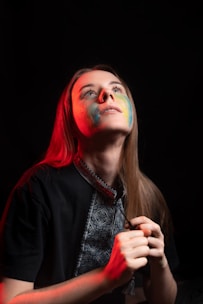 A person is looking upwards, with a serene expression. Their face is partially illuminated with colorful paint, primarily in blue and yellow, and they are bathed in dramatic red lighting from the side. The background is dark, enhancing the focus on the person. They are holding a piece of jewelry or clothing close to their chest.