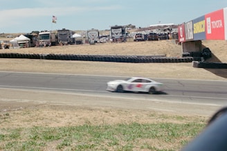 A racing car is captured in motion on a track with a blurred effect indicating high speed. Surrounding the track, there are RVs and tents where spectators are gathered, with various flags fluttering above. Advertising banners from different brands such as Toyota and Sunoco are displayed prominently on the bridge over the track.