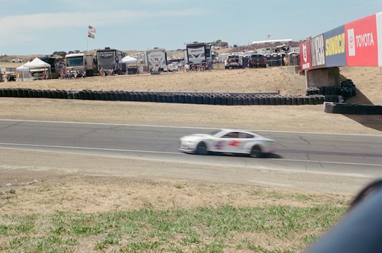 A racing car is captured in motion on a track with a blurred effect indicating high speed. Surrounding the track, there are RVs and tents where spectators are gathered, with various flags fluttering above. Advertising banners from different brands such as Toyota and Sunoco are displayed prominently on the bridge over the track.