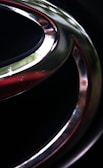 A close-up of a metallic emblem featuring a smooth, curved shape with reflective surfaces. The emblem is set against a dark background, and the metal surfaces exhibit a shiny finish, with hints of light reflecting off them. The foreground contains sleek silver and subtle red accents.