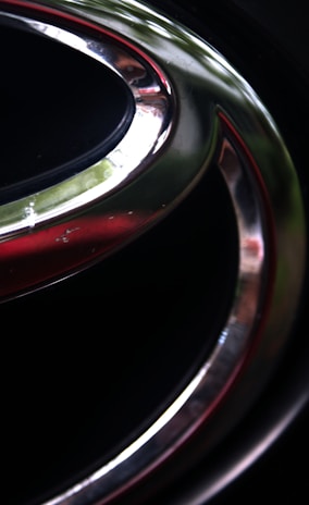 A close-up of a metallic emblem featuring a smooth, curved shape with reflective surfaces. The emblem is set against a dark background, and the metal surfaces exhibit a shiny finish, with hints of light reflecting off them. The foreground contains sleek silver and subtle red accents.