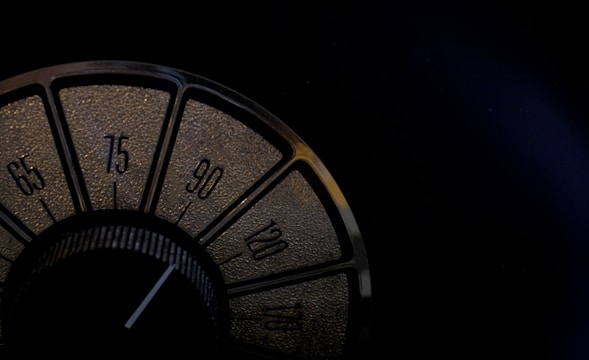 A close-up view of a vintage radio dial featuring numbers in increments of fifteen, from 65 to 120. The dial is set within a dark, textured background, and the numbers are highlighted in a golden hue.