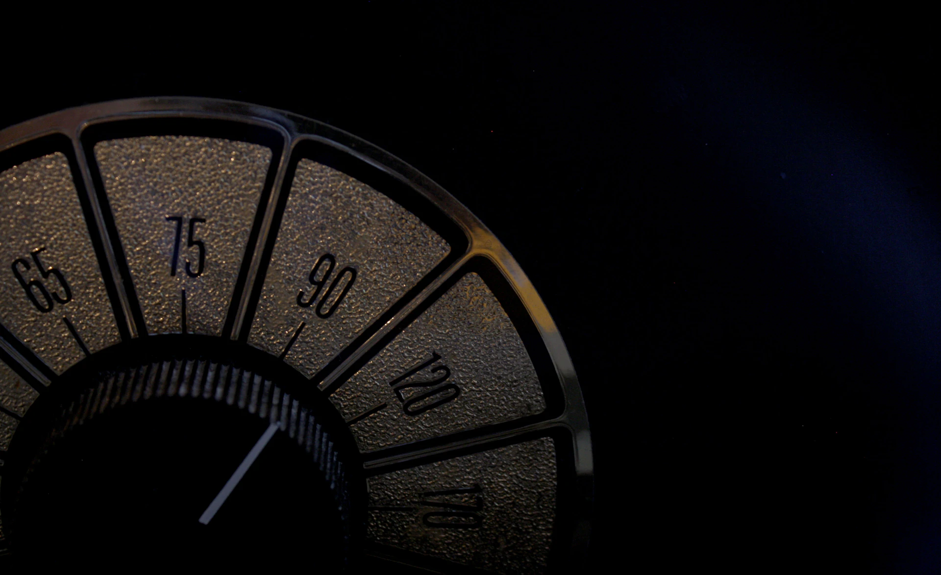 A close-up view of a vintage radio dial featuring numbers in increments of fifteen, from 65 to 120. The dial is set within a dark, textured background, and the numbers are highlighted in a golden hue.