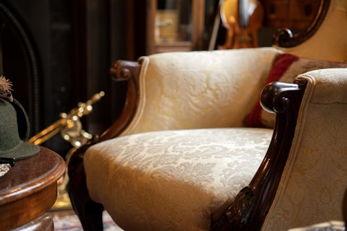 An elegant vintage armchair with ornately carved wooden arms and light patterned upholstery dominates a cozy room filled with classic decor. A green felt hat with a feather sits on a wooden table, while a brass fireplace tool rests in the background.