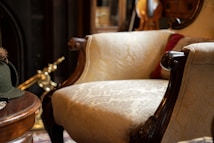 An elegant vintage armchair with ornately carved wooden arms and light patterned upholstery dominates a cozy room filled with classic decor. A green felt hat with a feather sits on a wooden table, while a brass fireplace tool rests in the background.