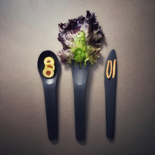 Three black utensils, consisting of a spoon, fork, and knife, are placed side by side. The spoon holds three slices of green olives, the fork is adorned with a leafy piece of lettuce, and two thin slices of ginger are positioned on the knife. The background is a neutral gray, enhancing the display of the simple, elegant arrangement.