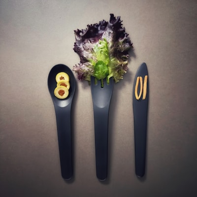 Three black utensils, consisting of a spoon, fork, and knife, are placed side by side. The spoon holds three slices of green olives, the fork is adorned with a leafy piece of lettuce, and two thin slices of ginger are positioned on the knife. The background is a neutral gray, enhancing the display of the simple, elegant arrangement.