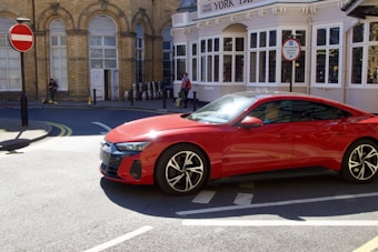A red sports car is parked on a street corner near a historic building with large windows and brickwork. A person is walking in the background, and there is a set of metal barrels lined up against the wall. Signs indicate an old pub named York Tap.