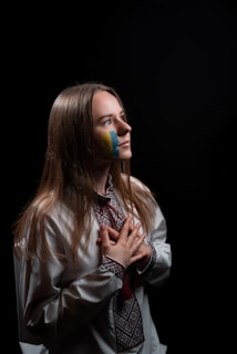 A young woman with long blonde hair stands against a dark background. She has a painted blue and yellow flag on her face and is dressed in traditional Eastern European attire. Her hands are clasped in front of her chest, and she gazes upwards with a solemn expression.