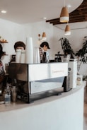 A cozy cafe interior features a large espresso machine on a curved white counter. Several paper cups are stacked on top, with a clear glass water bottle next to them. Two people are partially visible, one preparing a drink. Overhead, pendant lights hang from the ceiling, and there are wooden shelves in the background displaying small decorations. A large plant in the corner adds a touch of greenery.