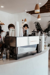 A cozy cafe interior features a large espresso machine on a curved white counter. Several paper cups are stacked on top, with a clear glass water bottle next to them. Two people are partially visible, one preparing a drink. Overhead, pendant lights hang from the ceiling, and there are wooden shelves in the background displaying small decorations. A large plant in the corner adds a touch of greenery.