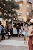 A lively outdoor scene featuring a vintage-style food truck with a sign reading 'CARAMEL'. People are gathered around, including adults and a child with a pink headband. The background consists of residential or commercial buildings with visible brickwork. A large tree provides shade over part of the area, and the atmosphere is relaxed and social.