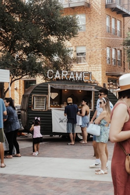 A lively outdoor scene featuring a vintage-style food truck with a sign reading 'CARAMEL'. People are gathered around, including adults and a child with a pink headband. The background consists of residential or commercial buildings with visible brickwork. A large tree provides shade over part of the area, and the atmosphere is relaxed and social.