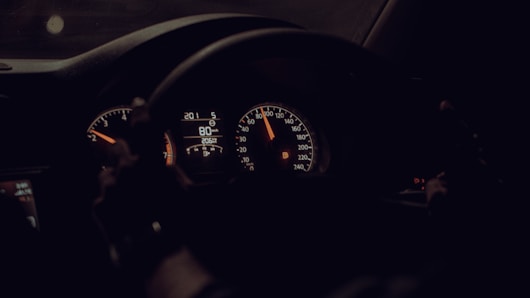 A dimly lit car dashboard with illuminated speedometers and gauges indicating a speed of 80 km/h. The steering wheel is visible, suggesting the driver's perspective.
