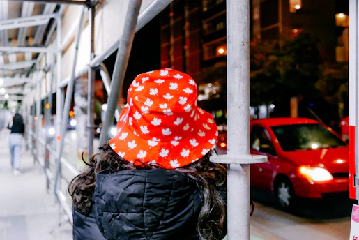 A person wearing a red bucket hat with white maple leaf patterns stands under scaffolding at night. In the background, there is a brightly lit street with a red car passing by.