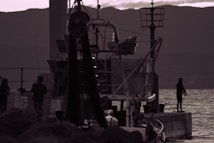 A silhouette of a fishing scene in low light with a dock extending into a body of water. There is a person fishing on the right side and several others near a boat or structure on the left. The background has mountains and a cloudy sky, while the overall tone is dark with a slight purple hue.
