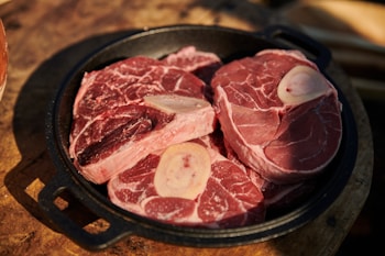 Several raw beef shank cuts with bone are placed inside a black cast iron skillet. The meat appears fresh with visible marbling, highlighting a rich red color. The skillet rests on a wooden surface, suggesting a rustic kitchen setting.