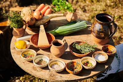 A rustic wooden table is set outdoors, displaying an assortment of fresh vegetables, herbs, and spices. Items include carrots, leeks, parsnips, and a lemon, as well as a wedge of cheese. Various small bowls contain herbs and spices, while a large jug and smaller cup add to the earthy, traditional aesthetic.