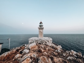 A lighthouse stands prominently atop a rocky cliff, surrounded by the vast expanse of the ocean. A clear sky stretches above, with a subtle moon visible, and a Greek flag flies on a flagpole in the foreground.