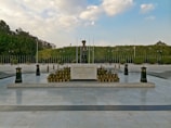 A ceremonial tombstone with inscriptions is surrounded by an array of small potted plants, set on a platform in a spacious outdoor area. The background features a well-manicured hedge and tall flagpoles, under a bright sky filled with scattered clouds.
