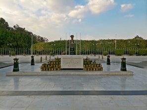 A ceremonial tombstone with inscriptions is surrounded by an array of small potted plants, set on a platform in a spacious outdoor area. The background features a well-manicured hedge and tall flagpoles, under a bright sky filled with scattered clouds.