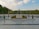 A ceremonial tombstone with inscriptions is surrounded by an array of small potted plants, set on a platform in a spacious outdoor area. The background features a well-manicured hedge and tall flagpoles, under a bright sky filled with scattered clouds.