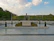 A ceremonial tombstone with inscriptions is surrounded by an array of small potted plants, set on a platform in a spacious outdoor area. The background features a well-manicured hedge and tall flagpoles, under a bright sky filled with scattered clouds.