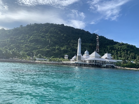 A serene coastal scene featuring a large, elegant white mosque with multiple domes and a tall minaret, set against a backdrop of lush green foliage on a hill. The foreground displays clear turquoise waters under a partly cloudy sky.