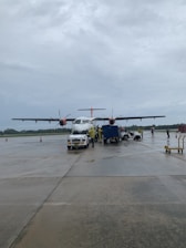 An airplane is parked on an airport tarmac with overcast skies. Ground crew members and service vehicles are positioned around the aircraft, and a few cones are visible nearby.