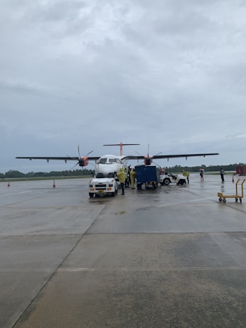 An airplane is parked on an airport tarmac with overcast skies. Ground crew members and service vehicles are positioned around the aircraft, and a few cones are visible nearby.