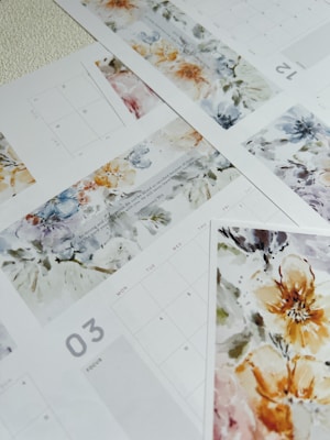 Multiple sheets of a calendar with watercolor floral designs. The calendar pages display different months, each adorned with intricate and colorful flower patterns. The overall design merges functionality with artistic aesthetics, blending calendar grids with vibrant artwork.