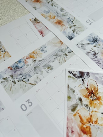 Multiple sheets of a calendar with watercolor floral designs. The calendar pages display different months, each adorned with intricate and colorful flower patterns. The overall design merges functionality with artistic aesthetics, blending calendar grids with vibrant artwork.