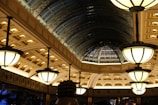 A lavishly designed interior space features a series of large, ornate light fixtures hanging from the ceiling. The architecture includes a series of arched windows with an intricate metal framework, creating a spacious, elegant atmosphere. The ceiling is adorned with detailed patterns and recessed lighting, casting a warm glow throughout the area.