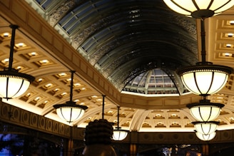 A lavishly designed interior space features a series of large, ornate light fixtures hanging from the ceiling. The architecture includes a series of arched windows with an intricate metal framework, creating a spacious, elegant atmosphere. The ceiling is adorned with detailed patterns and recessed lighting, casting a warm glow throughout the area.