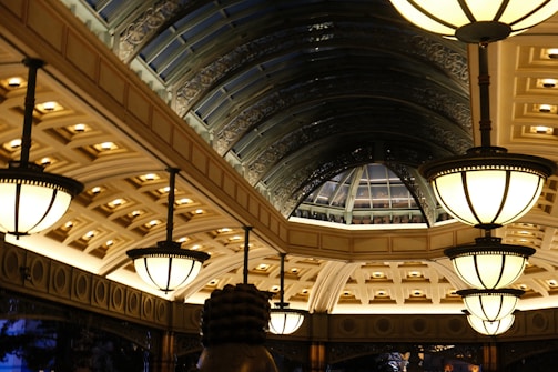 A lavishly designed interior space features a series of large, ornate light fixtures hanging from the ceiling. The architecture includes a series of arched windows with an intricate metal framework, creating a spacious, elegant atmosphere. The ceiling is adorned with detailed patterns and recessed lighting, casting a warm glow throughout the area.