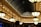 A lavishly designed interior space features a series of large, ornate light fixtures hanging from the ceiling. The architecture includes a series of arched windows with an intricate metal framework, creating a spacious, elegant atmosphere. The ceiling is adorned with detailed patterns and recessed lighting, casting a warm glow throughout the area.
