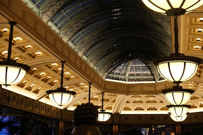 A lavishly designed interior space features a series of large, ornate light fixtures hanging from the ceiling. The architecture includes a series of arched windows with an intricate metal framework, creating a spacious, elegant atmosphere. The ceiling is adorned with detailed patterns and recessed lighting, casting a warm glow throughout the area.