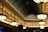 A lavishly designed interior space features a series of large, ornate light fixtures hanging from the ceiling. The architecture includes a series of arched windows with an intricate metal framework, creating a spacious, elegant atmosphere. The ceiling is adorned with detailed patterns and recessed lighting, casting a warm glow throughout the area.