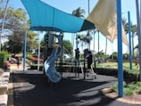 A playground with a blue plastic slide and climbing structure under a shade sail. Children are playing on the equipment. The area is surrounded by palm trees and greenery. A woman is walking on the pathway nearby.