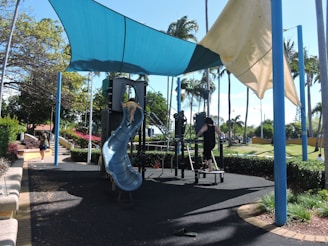A playground with a blue plastic slide and climbing structure under a shade sail. Children are playing on the equipment. The area is surrounded by palm trees and greenery. A woman is walking on the pathway nearby.