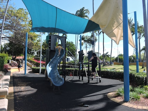 A playground with a blue plastic slide and climbing structure under a shade sail. Children are playing on the equipment. The area is surrounded by palm trees and greenery. A woman is walking on the pathway nearby.