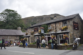 A quaint stone inn adorned with colorful flowers is set against a hilly backdrop. People are gathered around, some walking dogs while others are engaged in conversation. A motorcycle is parked nearby, adding a touch of modernity to the rustic scene. The atmosphere is lively, with a sense of community.